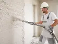 Budowlaniec w kasku i goglach nakłada tynk maszynowy na ścianę. Dowiedz się, ile kosztują tynki maszynowe.