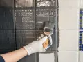 Ręka w białej rękawiczce maluje pędzlem szarą farbą płytki ceramiczne, szukając jaka najlepsza farba do płytek ceramicznych.