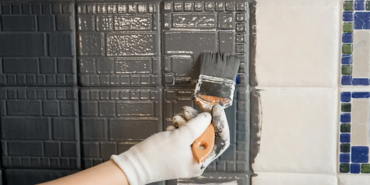 Ręka w białej rękawiczce maluje pędzlem szarą farbą płytki ceramiczne, szukając jaka najlepsza farba do płytek ceramicznych.