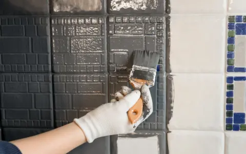 Ręka w białej rękawiczce maluje pędzlem szarą farbą płytki ceramiczne, szukając jaka najlepsza farba do płytek ceramicznych.