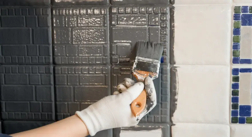 Ręka w białej rękawiczce maluje pędzlem szarą farbą płytki ceramiczne, szukając jaka najlepsza farba do płytek ceramicznych.